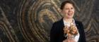 Archaeologist, Dr Susanna Harris standing in front of a swirly mosaic holding a bunch of round ceramic weights on sticks. These are replica spindles, tools used to twist fibres into yarn.