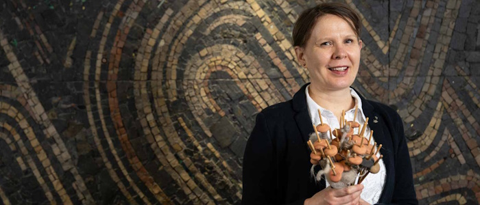 Archaeologist, Dr Susanna Harris standing in front of a swirly mosaic holding a bunch of round ceramic weights on sticks. These are replica spindles, tools used to twist fibres into yarn.