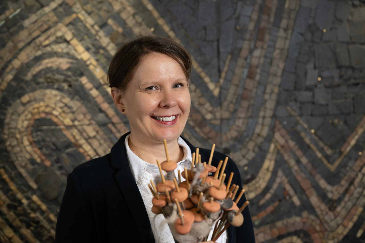 Archaeologist, Dr Susanna Harris standing in front of a swirly mosaic holding a bunch of round ceramic weights on sticks. These are replica spindles, tools used to twist fibres into yarn.