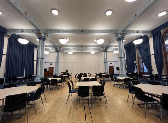 Flat floored hall with tables and chairs in groups, video monitors, lectern, and PC.