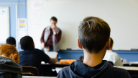 A photo taken from the back of classroom, showing the backs of students sitting at desks and the teach at the front of the room