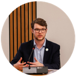 Daniel is sat at a desk speaking into a microphone. He is wearing a dark suit blue shirt. He has brown hair and wears glasses.