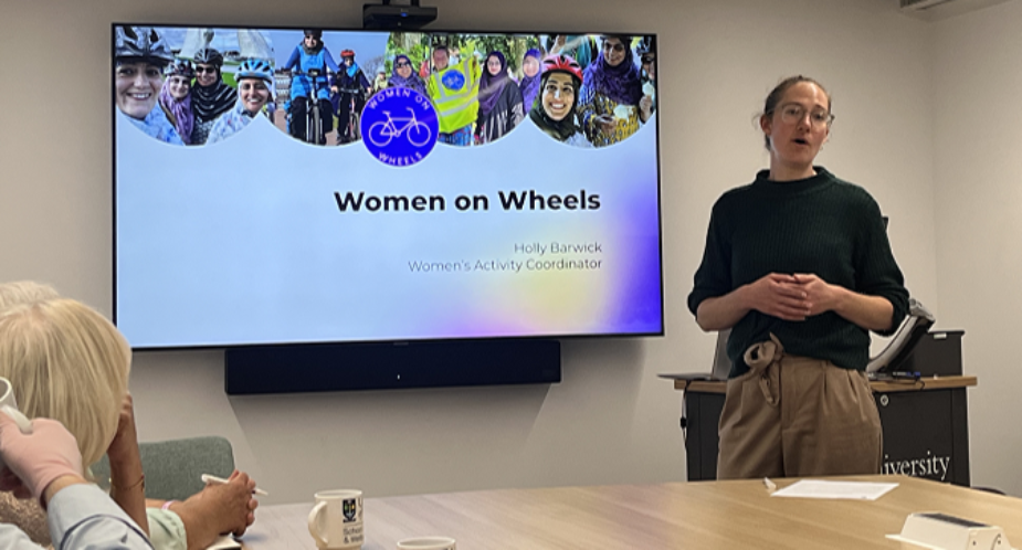 A woman is standing beside a screen talking about Women on Wheels