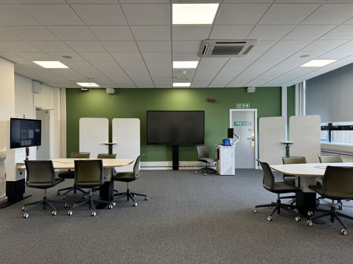 Flat floored teaching room with tables and chairs, each with a video monitor, moveable whiteboards, large video monitor, lectern, PC, and lecturer's chair.