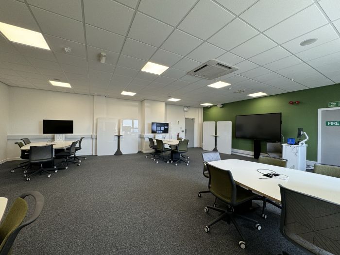 Flat floored teaching room with tables and chairs, each with a video monitor, moveable whiteboards, large video monitor, lectern, PC, and lecturer's chair.