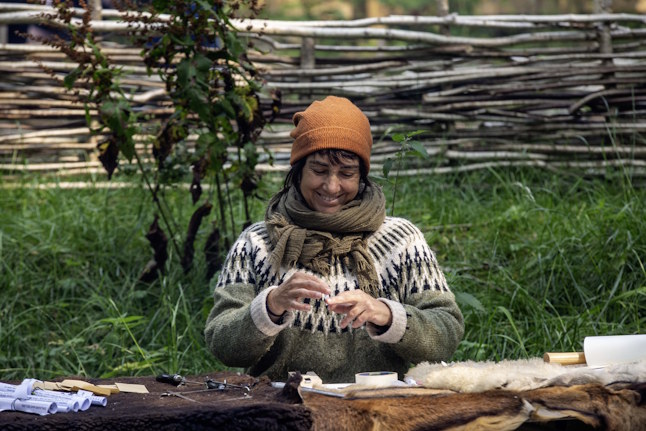 Nerea Bello sitting at a table covered with pelts rolling up a tiny strip of paper. There are tools for making jewellery on the table.