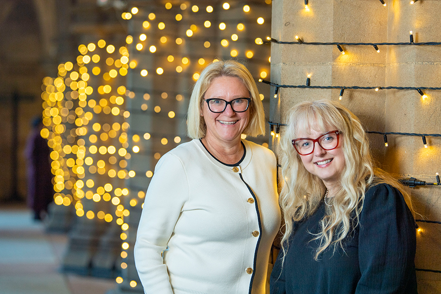 Two women with blonde hair, one in black dress and one in white blouse, smiling at the camera while standing by the cloisters with twinkling lights
