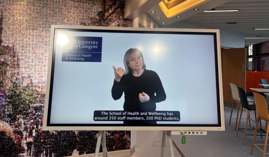 A large screen displays a woman using sign language. The text says - The School of Health and Wellbeing has around 350 staff members, 200 PhD students