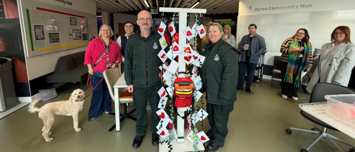 Two Scottish Ambulance Service staff members with a defibrillator. People and a dog are standing behind them.