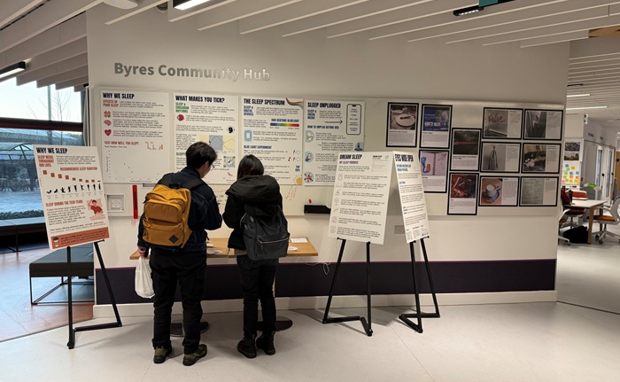 Two people are looking at the Eyes Wide Open adolescent sleep exhibition. There are posters on the wall and on display stands.
