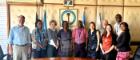 A group of ten people pose for a photo in a formal meeting room. They are standing in front of a wall displaying the logo of the Kenya Medical Research Institute (KEMRI), along with several national and institutional flags. The individuals represent a diverse team, with some wearing lanyards. A framed portrait hangs above the KEMRI logo.