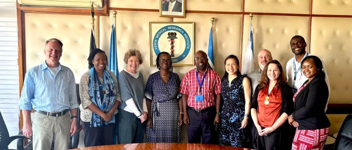A group of ten people pose for a photo in a formal meeting room. They are standing in front of a wall displaying the logo of the Kenya Medical Research Institute (KEMRI), along with several national and institutional flags. The individuals represent a diverse team, with some wearing lanyards. A framed portrait hangs above the KEMRI logo.