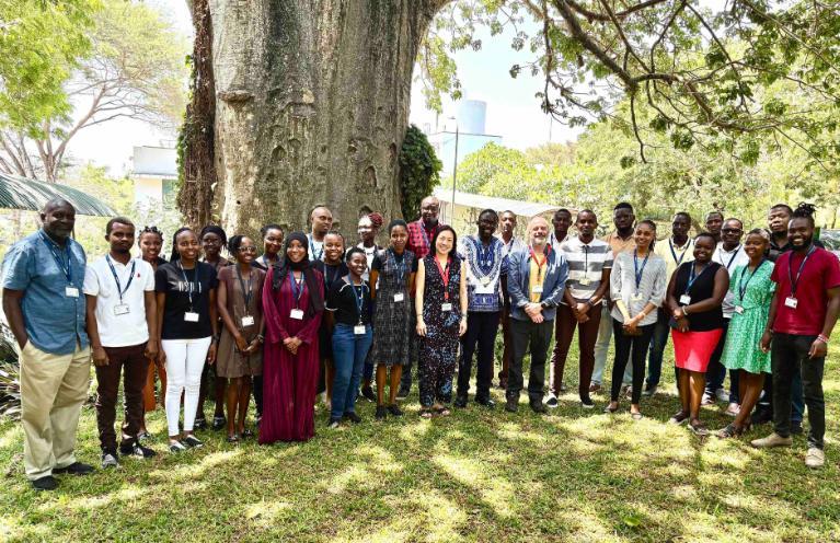 A large group of people poses outdoors under a massive tree with a textured trunk, surrounded by greenery. Many individuals are wearing lanyards, indicating a professional or academic gathering. The group is diverse, with a mix of ages, genders, and styles of dress, including casual and professional attire