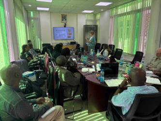 A group of professionals is seated around a large, dark wooden conference table in a well-lit meeting room. A presenter, a woman wearing a light green cardigan, stands at the front, gesturing towards a screen displaying a slide with text and images. The attendees, a diverse group, are engaged, taking notes, and using laptops.