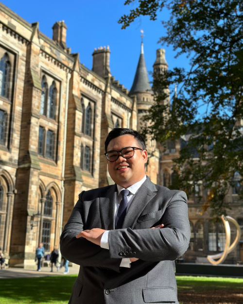 Albert Ghana Pratama wearing a suit, stood in front of the Gilbert Scott Building on a sunny day