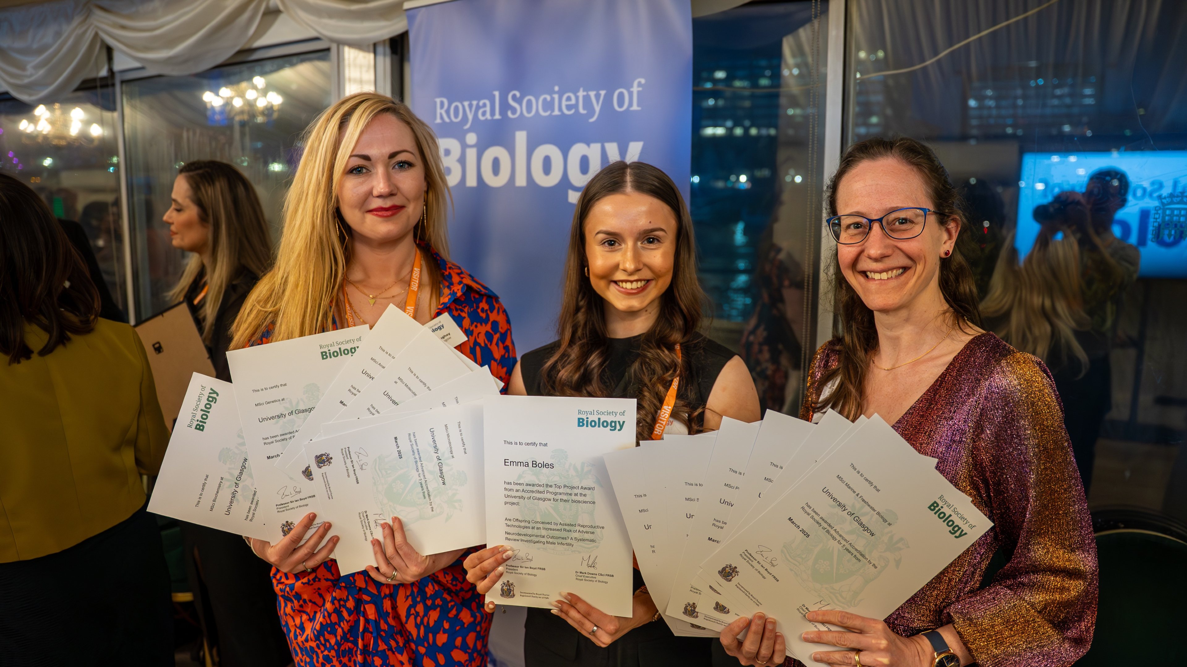 Dr Laura McCaughey, hons student Emma Boles, and Dr Anna McGregor stood in front of the RSB logo on a pull-up stand at the RSB ceremony. The trio are each holding certificates of the 16 accredited degrees