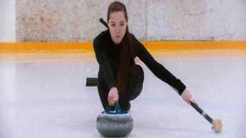 Woman in black athletic wear sliding a curling stone on an ice rink, preparing to release it.