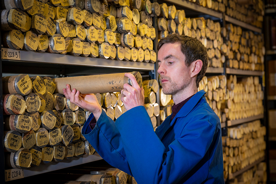 Michael Gallagher wearing a blue dust jacket, retrieving a manuscript in a cylinder from a shelf in the archives full of others