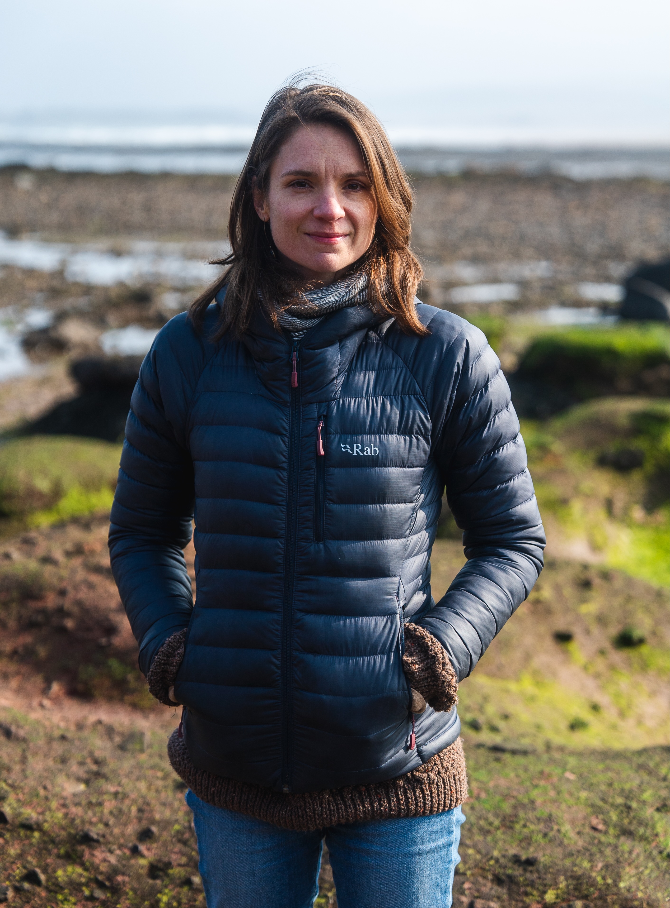Photo of a white woman with brown hair wearing a jacket on a rocky beach. Credit: Odin Gillies