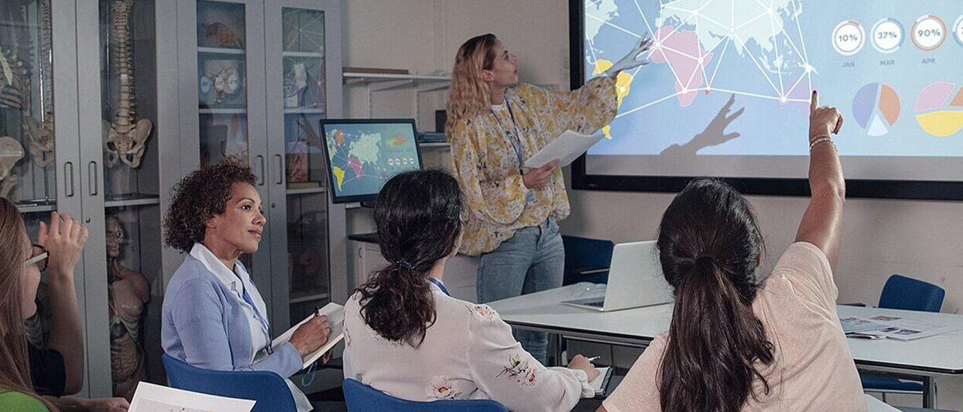 A female techer pointing at a projection with a class looking on