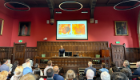 A wide-angle shot showing Professor Ho at the podium and the presentation screen, taken from further back in the room showing the engaged audience