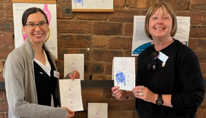 Dr Megan MacLeod and Jane Topping stood in front of an indoor brick wall, holding up paper to show the science-inspired art