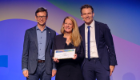 Dr Eve Smith stood on the stage at the EULAR Conference holding a certificate of her award, with those presenting the award stood on either side