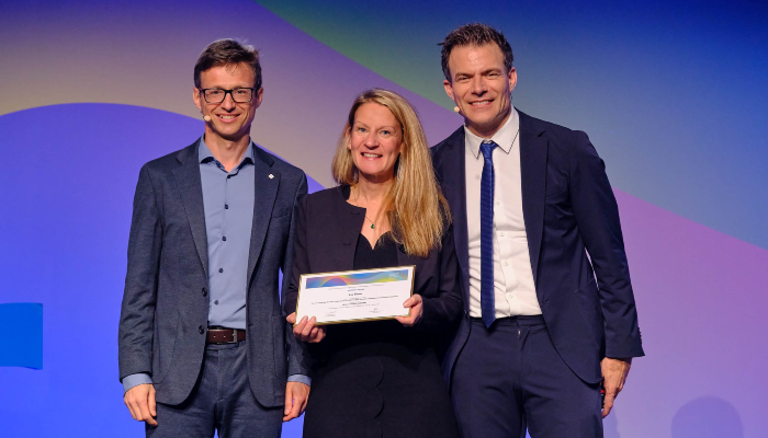 Dr Eve Smith stood on the stage at the EULAR Conference holding a certificate of her award, with those presenting the award stood on either side