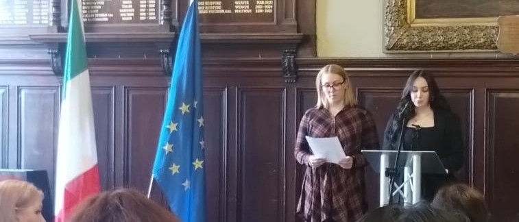 Carys Bilton and Eva Merritt stand behind a podium and give a reading to an assembled crowd. An Italian and an EU flag stand next to them