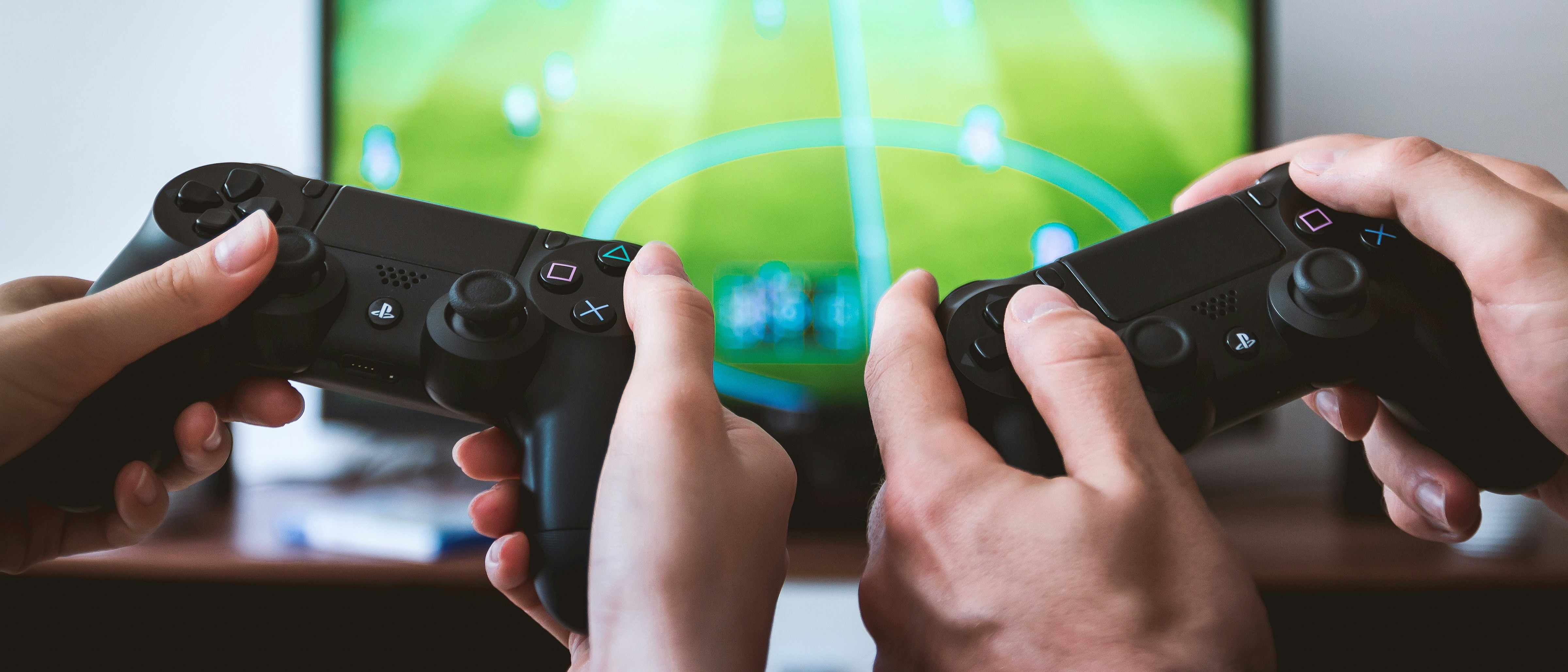 Two sets of hands hold PlayStation controllers while a video game is visible in the background.