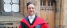 Professor Stephen Forcer stands in the cloisters wearing academic dress.