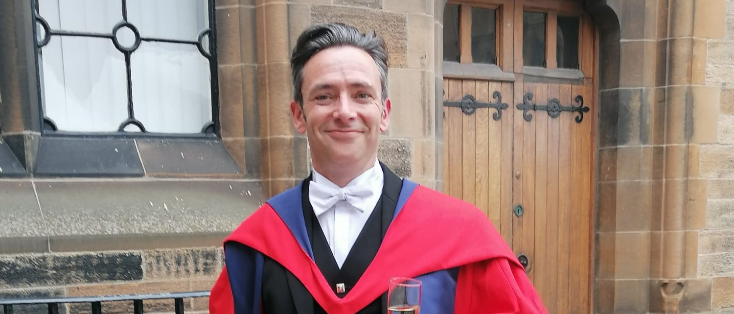 Professor Stephen Forcer stands in the cloisters wearing academic dress.