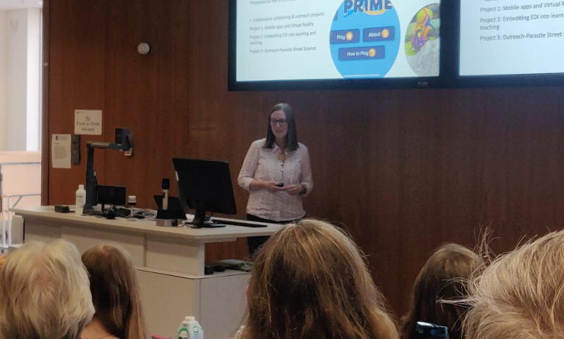 Professor Nicola Veitch delivering her inaugural lecture. She is stood in the wood-panelled lecture theatre behind the podium and directly below the double screen mounted on the wall, showing her presentation slides..