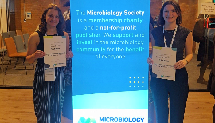 CVR PhD student Emma Davies stood left of a bright blue Microbiology Society banner with her Early Career Microbiologist of the Year certificate, with Microbiology PhD student Phoebe Crossley stood right of the banner with her third place certificate.
