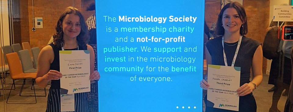 CVR PhD student Emma Davies stood left of a bright blue Microbiology Society banner with her Early Career Microbiologist of the Year certificate, with Microbiology PhD student Phoebe Crossley stood right of the banner with her third place certificate.