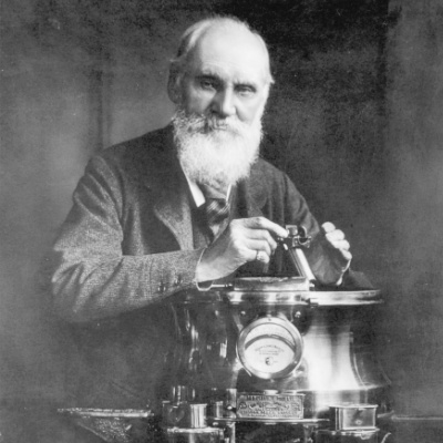 Photo of Lord Kelvin wearing a suit, looking to camera, positioned behind a piece of equipment