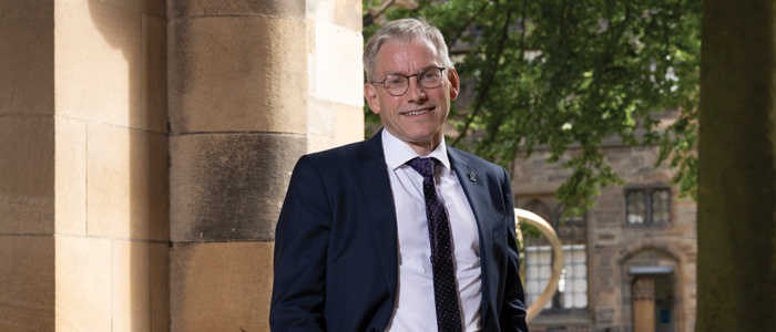 Prof Andy Schofield standing in the cloisters wearing a navy suit and smiling to the camera