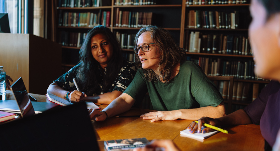 Katie sat at a table n a library with three colleagues - one is clearly in vision - and Katie is pointing at a laptop