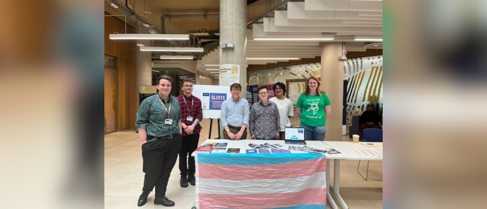 6 students standing at a desk with a transgender flag hanging from the front of the table. Leaflets and fliers are on the table with a board with information about GLINTS behind.