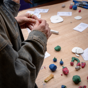 Someone holding some clay and molding it into a food shape. In the background you can see a table with pieces of colourful clay and cards laying on it.