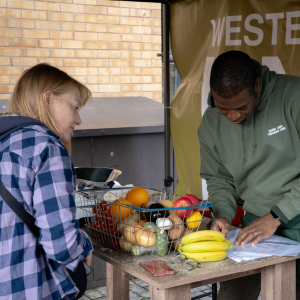 A member of staff checking the price list while they serve a customer who has a basket of veg placed on the counter.