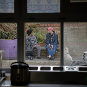 Two people pictured through a window. They are sitting on a wall outside talking.
