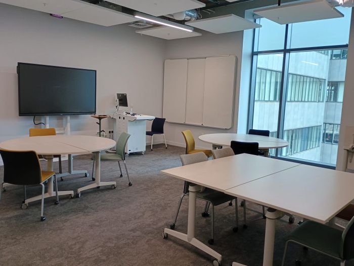 Flat floored teaching room with tables and chairs, PC, lectern, lecturer's chair, whiteboard, and video monitor.