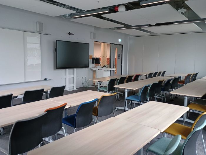 Flat floored teaching room with tables and chairs, PC, lectern, whiteboard, and video monitor.