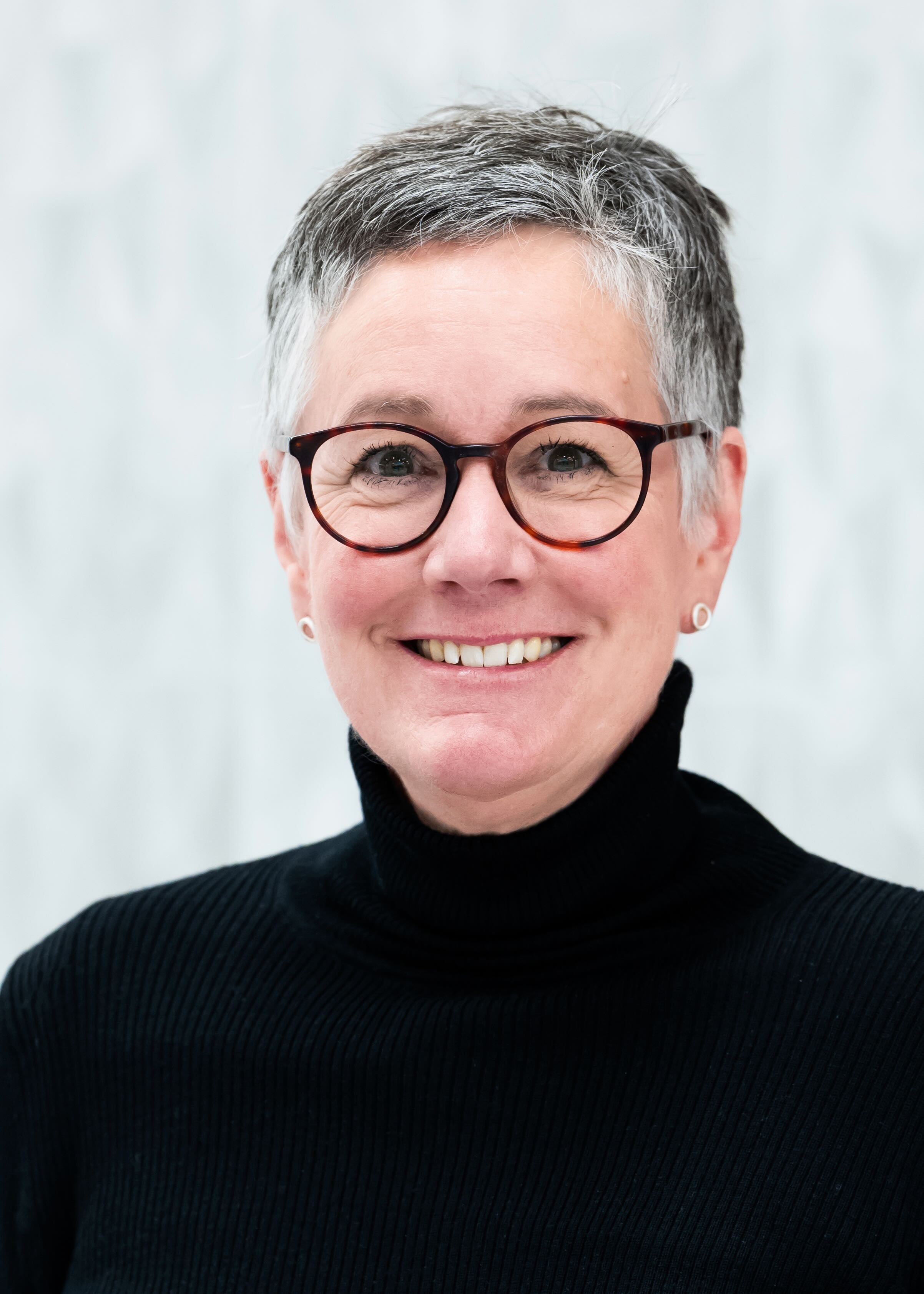 Person wearing a black ribbed turtleneck sweater, with short gray hair styled in a neat, slightly wavy cut, and thin-framed glasses, standing against a light gray textured background
