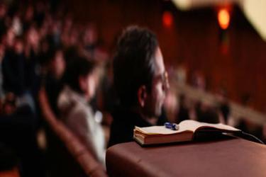 A photo of a lecture hall filled with people, a man in the foreground watches with a book and pen next to him