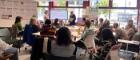 A group of people sitting round a big table in Byres Community Hub. They are listening to a speaker who is presenting information on a screen