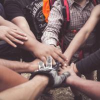 Several people, standing in a circle, are putting their hands together in the middle, we mainly just see the hands.