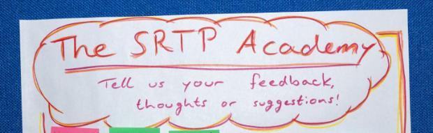 Handwritten sign reading ‘The SRTP Academy’ with the message ‘Tell us your feedback, thoughts or suggestions!’, written in red and pink marker on white paper against a blue background