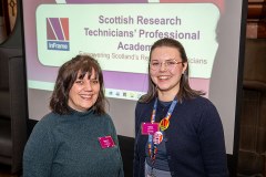 Two SRTP Academy representatives standing in front of a presentation slide reading ‘Scottish Research Technicians’ Professional Academy Symposium,’ wearing conference lanyards and smiling at the camera